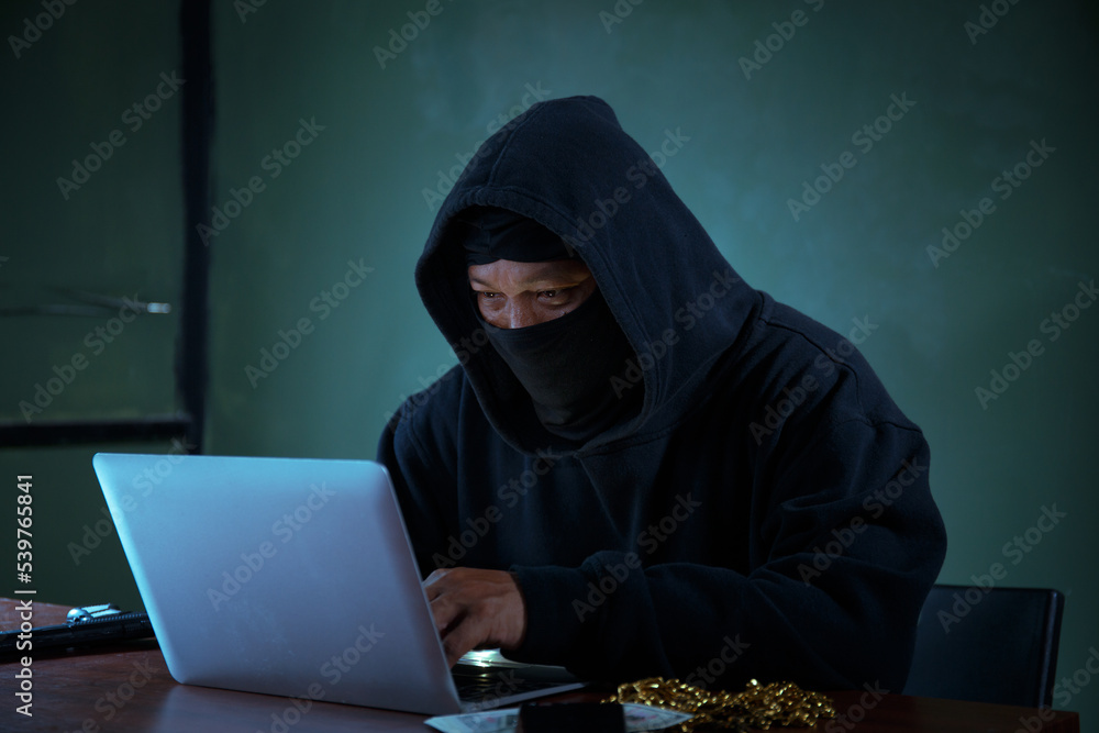 A male spy hacker in a black hoodie sits on a table in a dark room. looking at laptop computer A male spy hacker in a black hoodie sits on a table in a dark room. looking at laptop computer