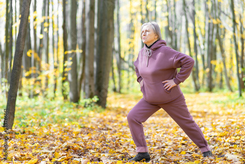 Older woman in a purple tracksuit is doing yoga outdoors. Performs exercises in the autumn park. Full length portrait.