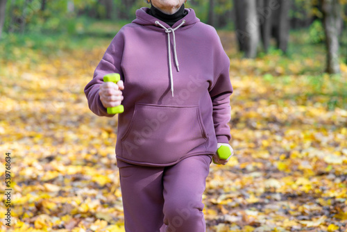 Unrecognizable pensioner woman performs exercises with small dumbbells in the park. Running with dumbbells in place. Outdoor fitness classes.