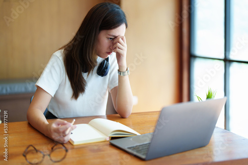 A portrait of a young Asian woman using a computer, wearing headphones and using a notebook to study online shows boredom and pain from video conferencing on a wooden desk in library © Jirapong