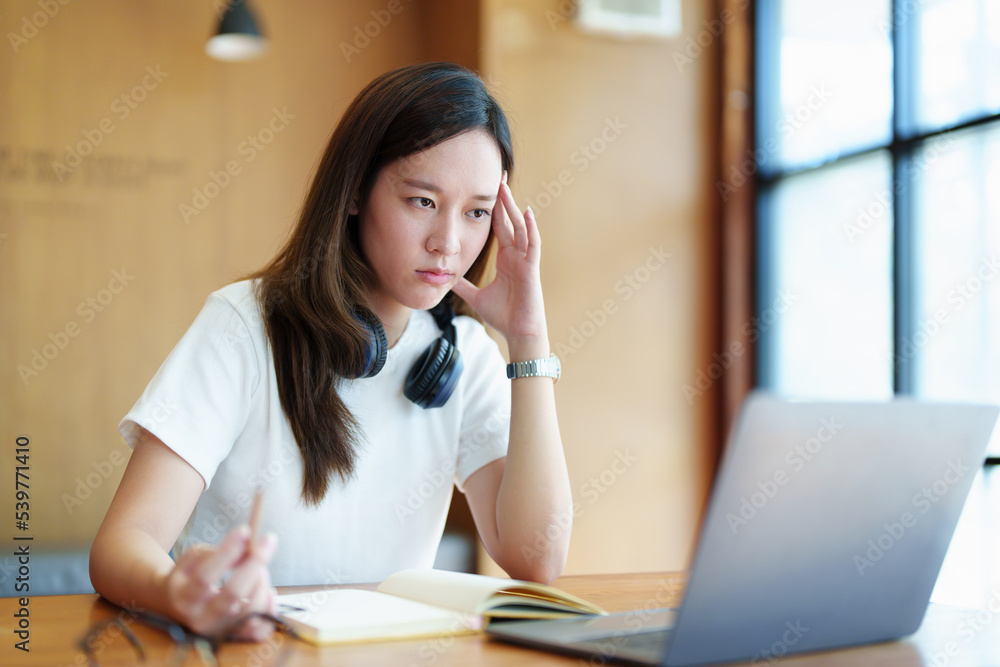 A portrait of a young Asian woman using a computer, wearing headphones and using a notebook to study online shows boredom and pain from video conferencing on a wooden desk in library A portrait of a young Asian woman using a computer, wearing headphones and using a notebook to study online shows boredom and pain from video conferencing on a wooden desk in library