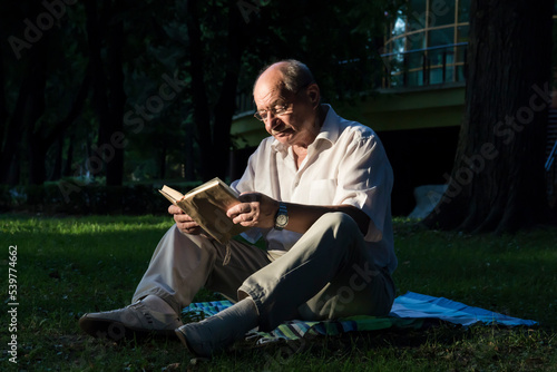 Fototapeta Naklejka Na Ścianę i Meble -  An elderly man in a white shirt is sitting on a blanket, on the ground in a park and reading an interesting book. A pensioner alone is resting in nature, passionate about his hobby.