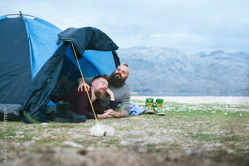 © Robert Petrovic - Attractive male gay couple camping by the sea under amazing mountain view. Hipsters homosexuals enjoying tenderness moment in a tent on empty beach. LGBT vacation in Autumn.