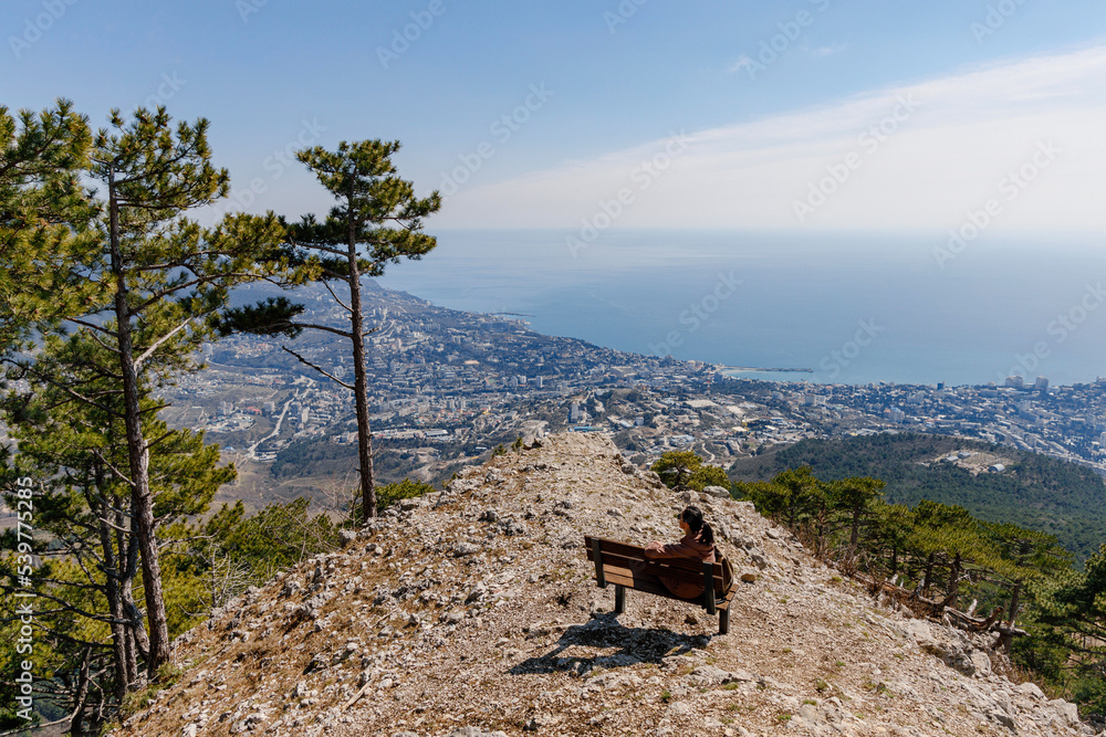 Woman sitting on a wooden bench on top of a hill with sky and city in the background on a sunny day