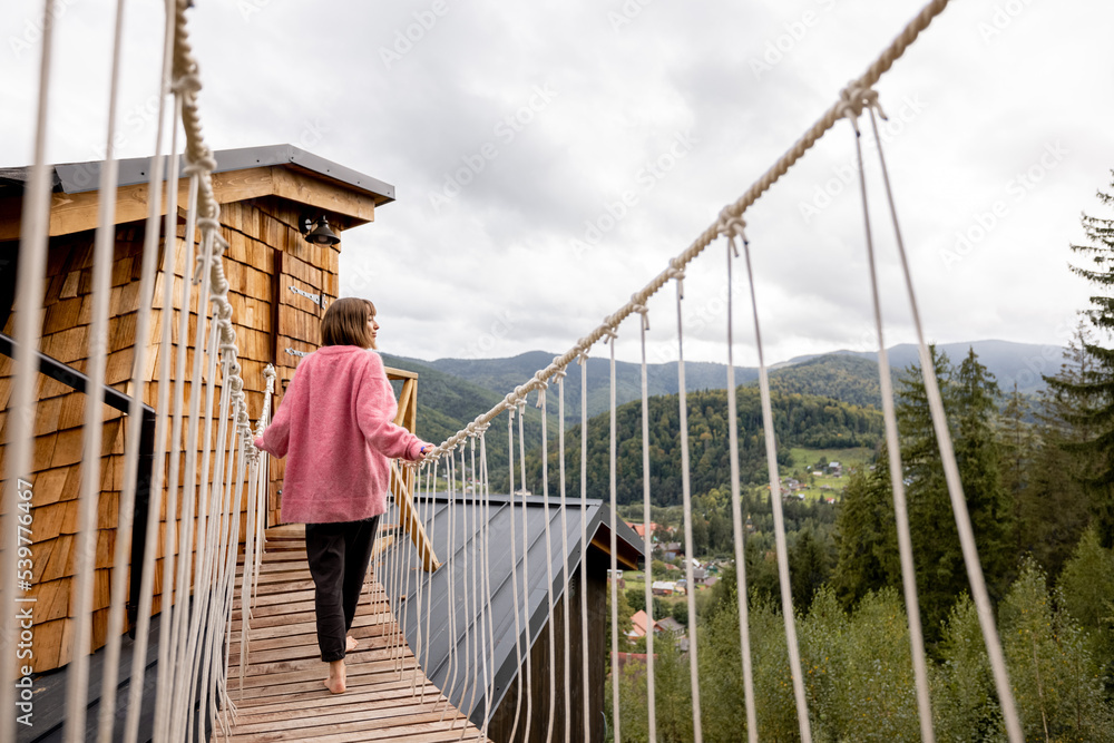 Woman enjoys scenic view on nature, while standing on the rope bridge of wooden house in mountains. Recreation and escaping to nature concept