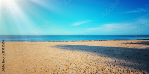 Fototapeta Naklejka Na Ścianę i Meble -  Bright long sunrays illuminate empty sandy beach with calm sea and clear blue sky