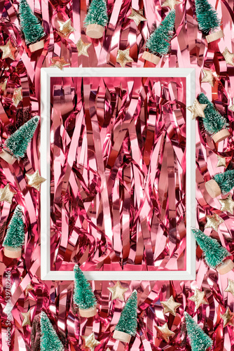 Toy Christmas trees and golden stars are scattered around a white empty photo frame on a shiny tinsel. Festive template for postcard, banner, Merry Christmas and Happy New Year greetings. Copy space.