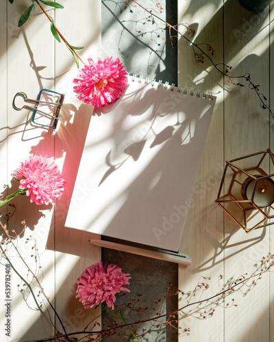 Flowers and an open empty notebook are on a wooden background, brightly lit by the light from the window. Nearby are stationery and dry branches of plants. Autumn floral arrangement with copy space.