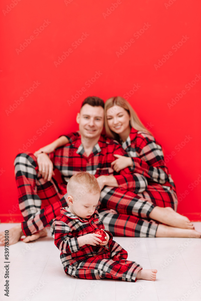 Happy family is having fun together. Smiling young mom, dad and baby son in red checked pyjamas sitting on red background. Mothers day. fathers day, baby day. Christmas. Valentines day