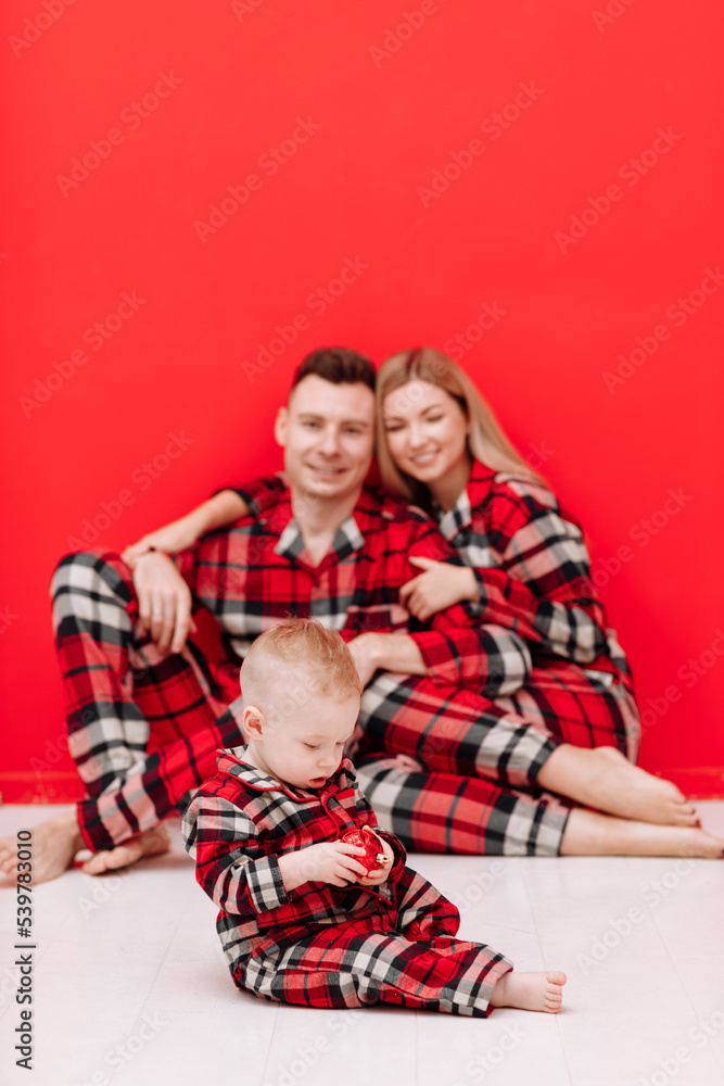 Happy family is having fun together. Smiling young mom, dad and baby son in red checked pyjamas sitting on red background. Mothers day. fathers day, baby day. Christmas. Valentines day Happy family is having fun together. Smiling young mom, dad and baby son in red checked pyjamas sitting on red background. Mothers day. fathers day, baby day. Christmas. Valentines day