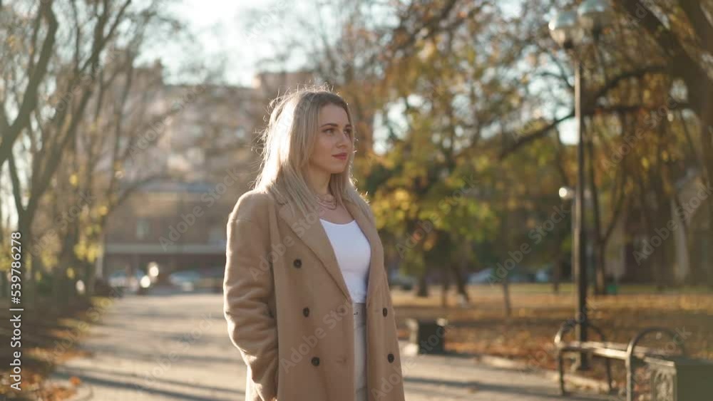 Portrait, Beautiful Business Woman Looks and Smiles at the Camera while Standing in an Autumn City Park on a Bright Sunny Day. Attractive Blonde in a Brown Coat. Positive Female Emotions. Slow Motion Portrait, Beautiful Business Woman Looks and Smiles at the Camera while Standing in an Autumn City Park on a Bright Sunny Day. Attractive Blonde in a Brown Coat. Positive Female Emotions. Slow Motion