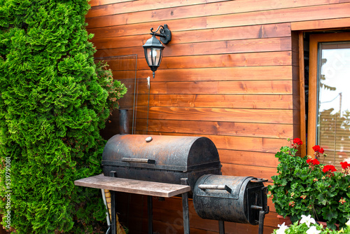 iron black rusty barbecue near wooden house with retro lantern on wall made of boards, cottage with thuja bushes and red pelargonium flowers, place to relax and cook meat and vegetables on grill.