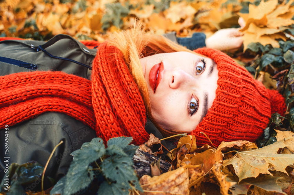 Autumn portrait of a cute girl in an orange warm knitted hat and an orange scarf lying in a fallen yellow leaf in the park. Romantic look of big eyes. Looks into the camera close-up.