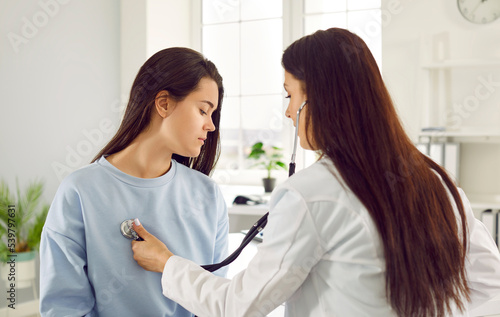 Young distressed Caucasian woman being examined in private clinic by female doctor with stethoscope to receive recommendations for treatment after illness sits in office of modern hospital