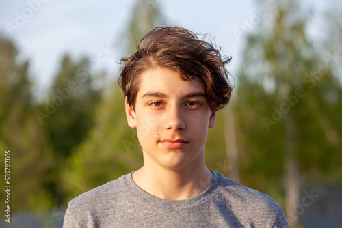 Portrait of a teenager in a grey T-shirt against a background of green foliage. Boy with brown eyes, lit by the setting sun, close-up © Klever_ok