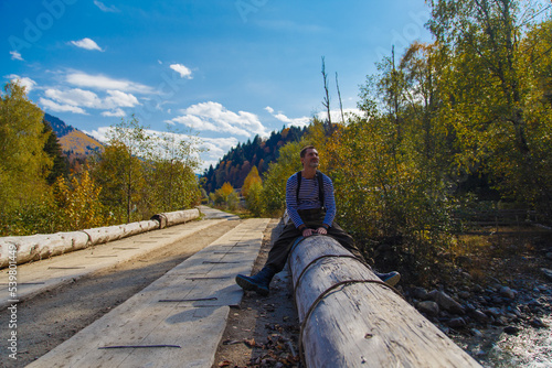 Portrait of a relaxed mature man sitting on a wooden log by a mountain river. A mature man enjoys a sunny autumn day by the river.