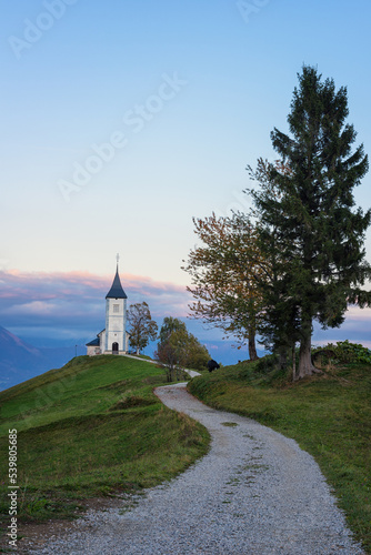 View of the Jamnik church St Primus and Felician at sunset, Alps mountains, Slovenia. Beautiful landscape with footpath, trees and blue sky with clouds, outdoor travel background