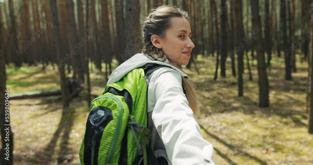 Delighted young blonde girl with braids smiling wearing backpack holding woman's hand heading through wood hikking trekking with mother. Women having fun resting.