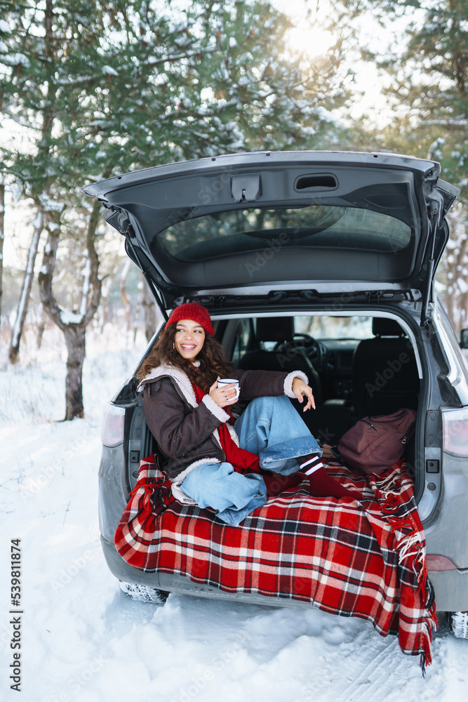Young woman holds a thermos and drinks tea sitting in car trunk in sunny winter forest. Rest, relaxation, travel, lifestyle concept.