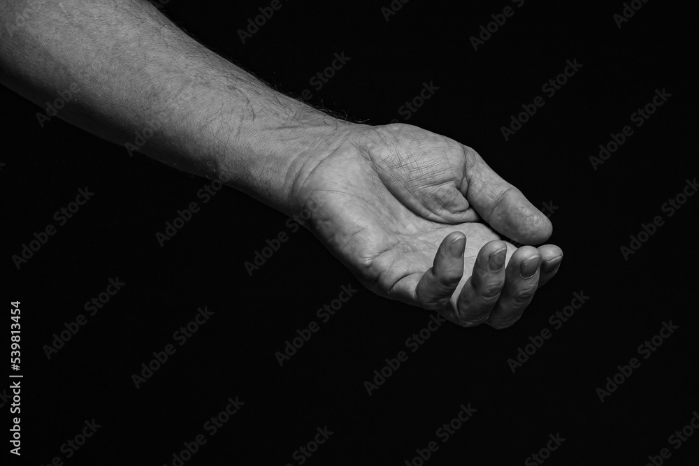 The outstretched wrinkled left hand of an elderly man asks for help on a black background. Black and white. The outstretched wrinkled left hand of an elderly man asks for help on a black background. Black and white.