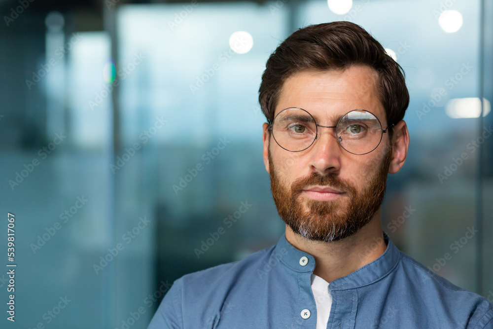 Close-up portrait of serious and thoughtful mature businessman with beard, man in shirt and glasses looking at camera, startuper working inside modern office building. Close-up portrait of serious and thoughtful mature businessman with beard, man in shirt and glasses looking at camera, startuper working inside modern office building.