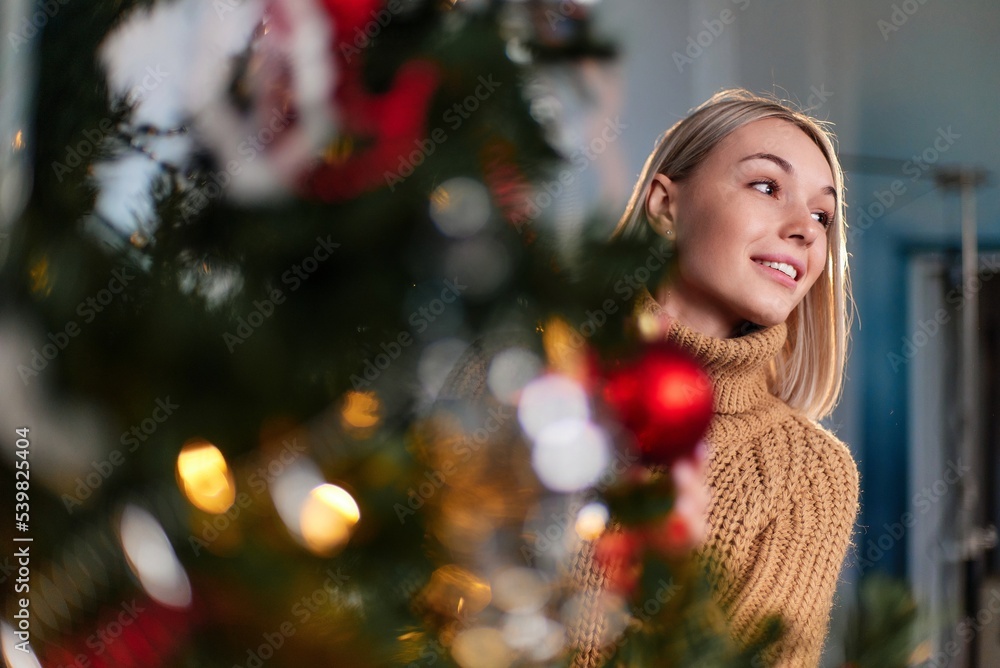 A beautiful girl peeks out from under the Christmas tree. Portrait of a young girl with Christmas trees. A beautiful girl in a knitted sweater next to a decorated Christmas tree. A beautiful girl peeks out from under the Christmas tree. Portrait of a young girl with Christmas trees. A beautiful girl in a knitted sweater next to a decorated Christmas tree.