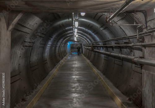 Long underground mountain tunnel with entrance in sight, corrugated metal walls, concrete floors, piping, nobody