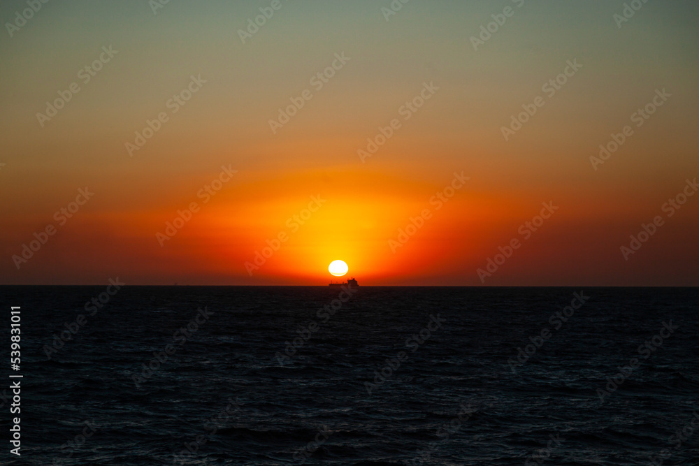 Sunset at sea and the silhouette of a transport ship in the distance on the horizon under the sun.