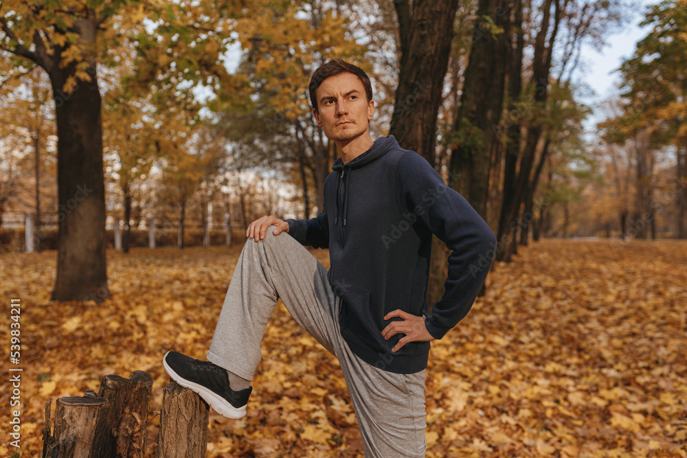 Fototapeta premium Confident sporty male athlete stretching legs leaning on tree while having fitness training outdoors in autumn park 