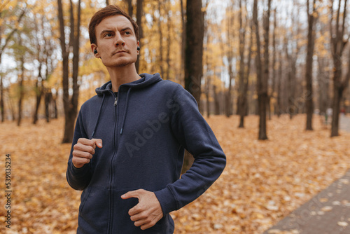 Fototapeta Naklejka Na Ścianę i Meble -  Confident male athlete in activewear running in autumn park and listening to music in wireless earbuds during outdoor training while leading healthy lifestyle  