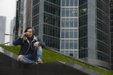 Young man with dreadlocks drinking coffee on background of skyscrapers. Relaxed hipster enjoying hot drink, sitting on stairway in downtown.