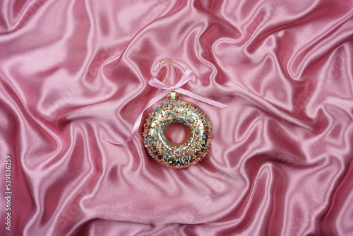 On a pink silk background is a Christmas tree toy in the form of a sweet donut. The concept of the new year. The concept of weight loss. The concept of a diet.