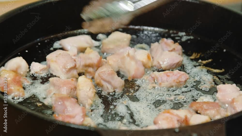 The cook stirs meat with culinary tongs, which is fried in boiling olive oil in a cast-iron frying pan The cook stirs meat with culinary tongs, which is fried in boiling olive oil in a cast-iron frying pan