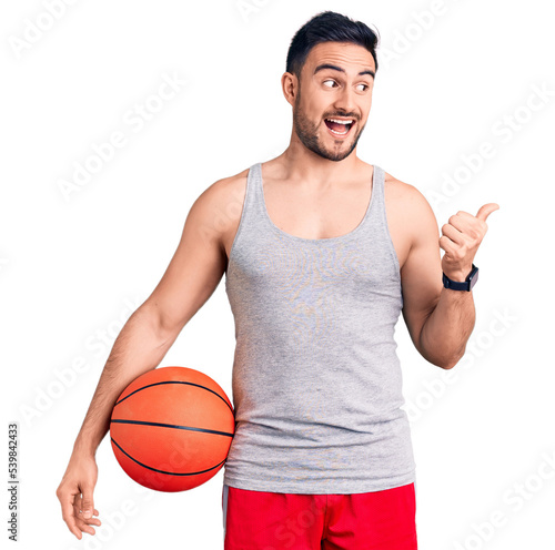 Young handsome man holding basketball ball pointing thumb up to the side smiling happy with open mouth