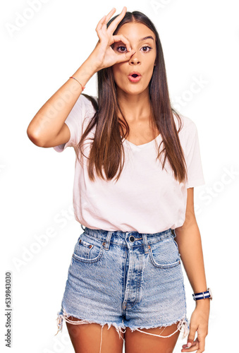 Young hispanic woman wearing casual white tshirt doing ok gesture shocked with surprised face, eye looking through fingers. unbelieving expression.