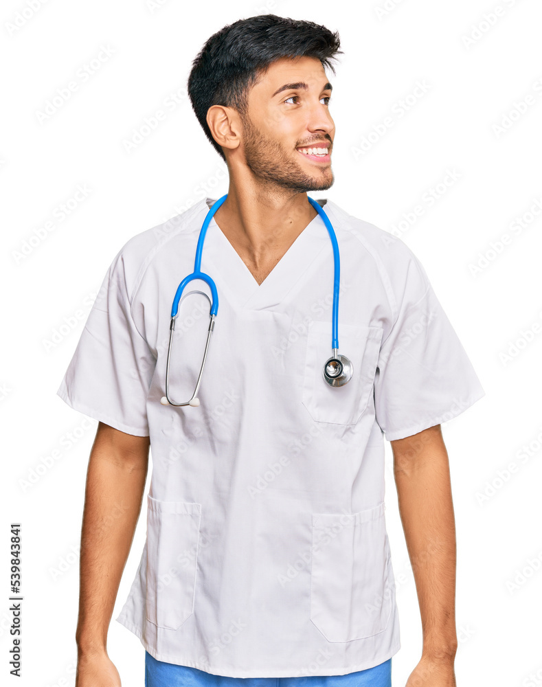 Young handsome man wearing doctor uniform and stethoscope looking away to side with smile on face, natural expression. laughing confident. Young handsome man wearing doctor uniform and stethoscope looking away to side with smile on face, natural expression. laughing confident.