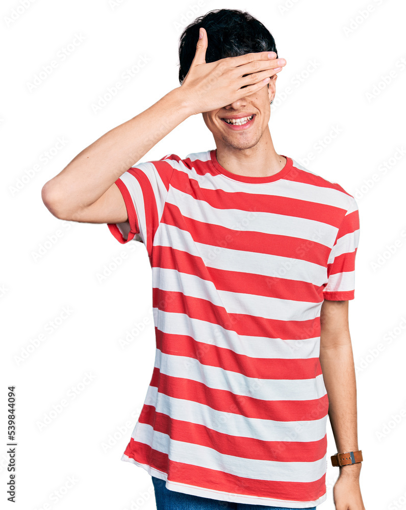Handsome hipster young man wearing casual striped t shirt smiling and laughing with hand on face covering eyes for surprise. blind concept. Handsome hipster young man wearing casual striped t shirt smiling and laughing with hand on face covering eyes for surprise. blind concept.