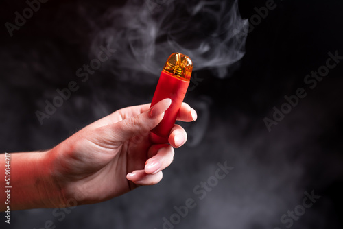 Red color vape pod system with replaceable cartridges in female hands isolated on black background. The concept of modern smoking, vaping and nicotine. Against the background of smoke