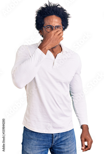 Handsome african american man with afro hair wearing casual clothes and glasses smelling something stinky and disgusting, intolerable smell, holding breath with fingers on nose. bad smell