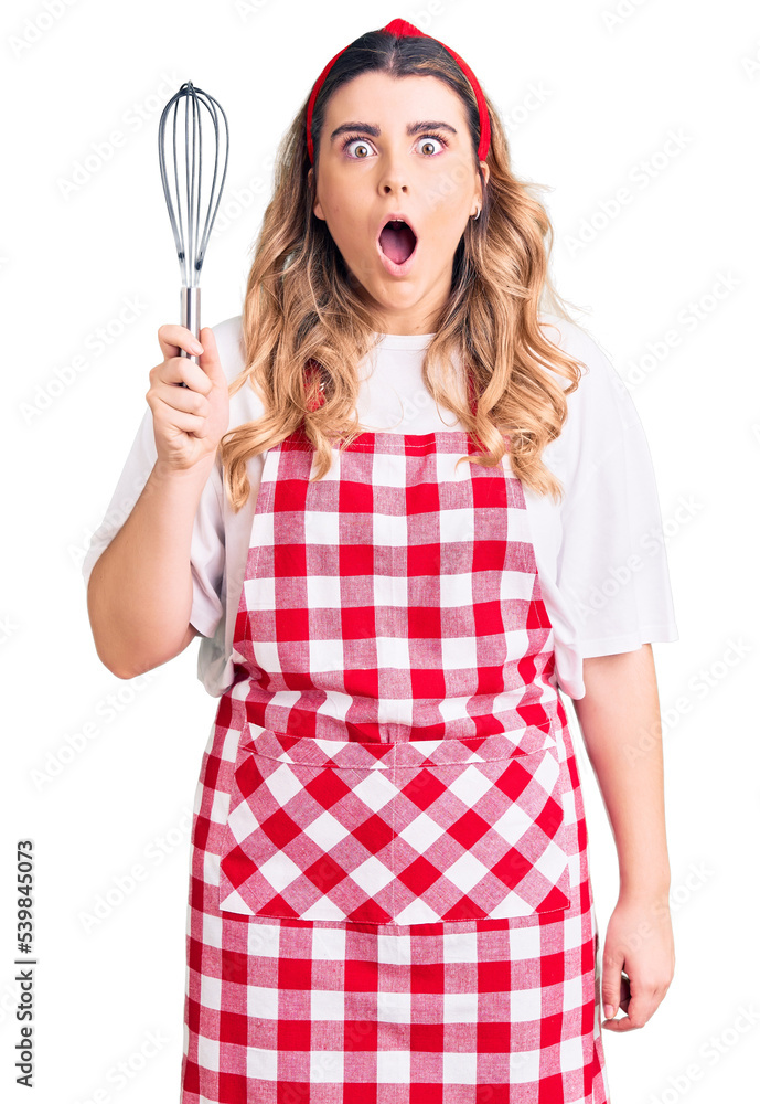 Young caucasian woman wearing apron holding whisk scared and amazed with open mouth for surprise, disbelief face Young caucasian woman wearing apron holding whisk scared and amazed with open mouth for surprise, disbelief face