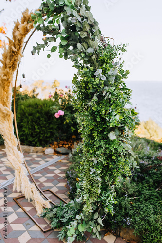 A round wedding arch  decorated with reeds  green leaves of plants  with electric hanging lamps  garlands  stands on a tile in the park  against the backdrop of the sea. Close-up photo.