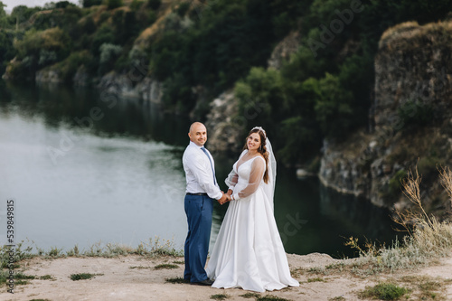 An adult groom and a beautiful, sweet bride in a white dress stand holding hands in nature near rocks, mountain cliffs against the backdrop of the sea. Wedding photography, portrait.
