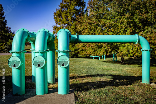 Teal colored pipes coming out of the ground in a park lawn. Their purpose is labeled as Conductivity, Temperature and Flow.