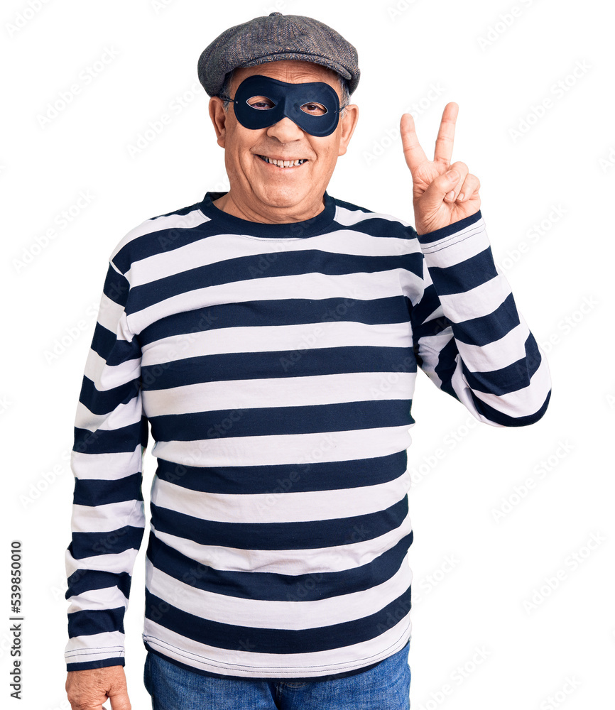Senior handsome man wearing burglar mask and t-shirt smiling with happy face winking at the camera doing victory sign with fingers. number two. Senior handsome man wearing burglar mask and t-shirt smiling with happy face winking at the camera doing victory sign with fingers. number two.