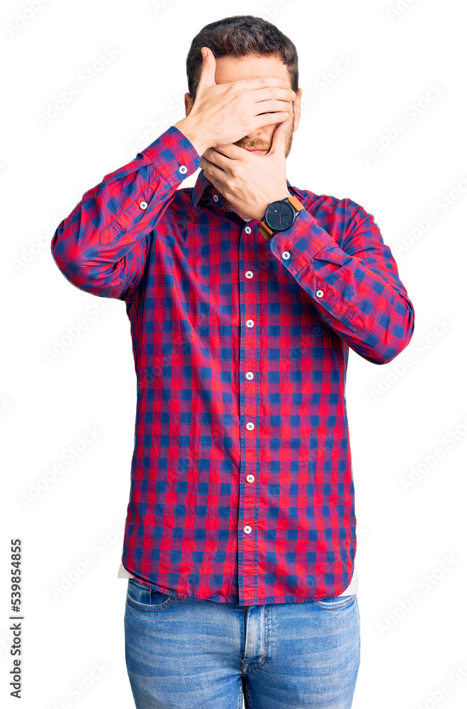 Handsome young man with bear wearing casual shirt covering eyes and mouth with hands, surprised and shocked. hiding emotion Handsome young man with bear wearing casual shirt covering eyes and mouth with hands, surprised and shocked. hiding emotion
