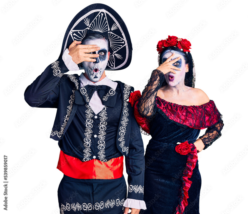 Young couple wearing mexican day of the dead costume over background peeking in shock covering face and eyes with hand, looking through fingers with embarrassed expression. Young couple wearing mexican day of the dead costume over background peeking in shock covering face and eyes with hand, looking through fingers with embarrassed expression.