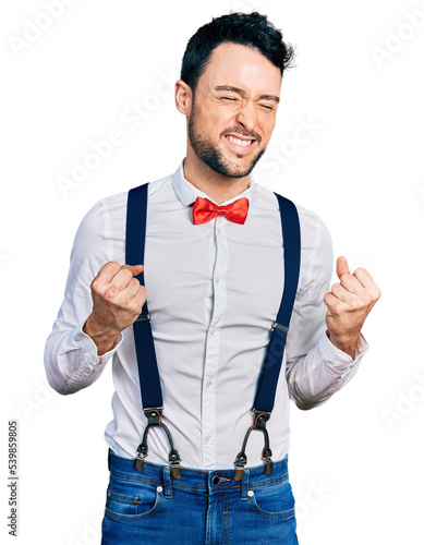 Hispanic man with beard wearing hipster look with bow tie and suspenders excited for success with arms raised and eyes closed celebrating victory smiling. winner concept.