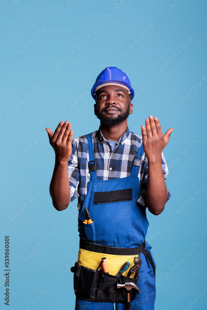 African american builder wearing coveralls and hard hat praying to God for new construction contracts. Builder in studio shot against blue background concept renovations and construction improvements. African american builder wearing coveralls and hard hat praying to God for new construction contracts. Builder in studio shot against blue background concept renovations and construction improvements.