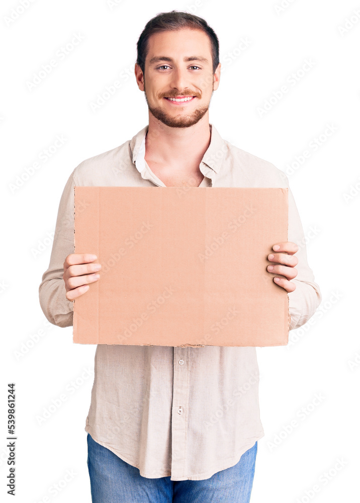 Young handsome caucasian man holding empty cardboard banner looking positive and happy standing and smiling with a confident smile showing teeth Young handsome caucasian man holding empty cardboard banner looking positive and happy standing and smiling with a confident smile showing teeth