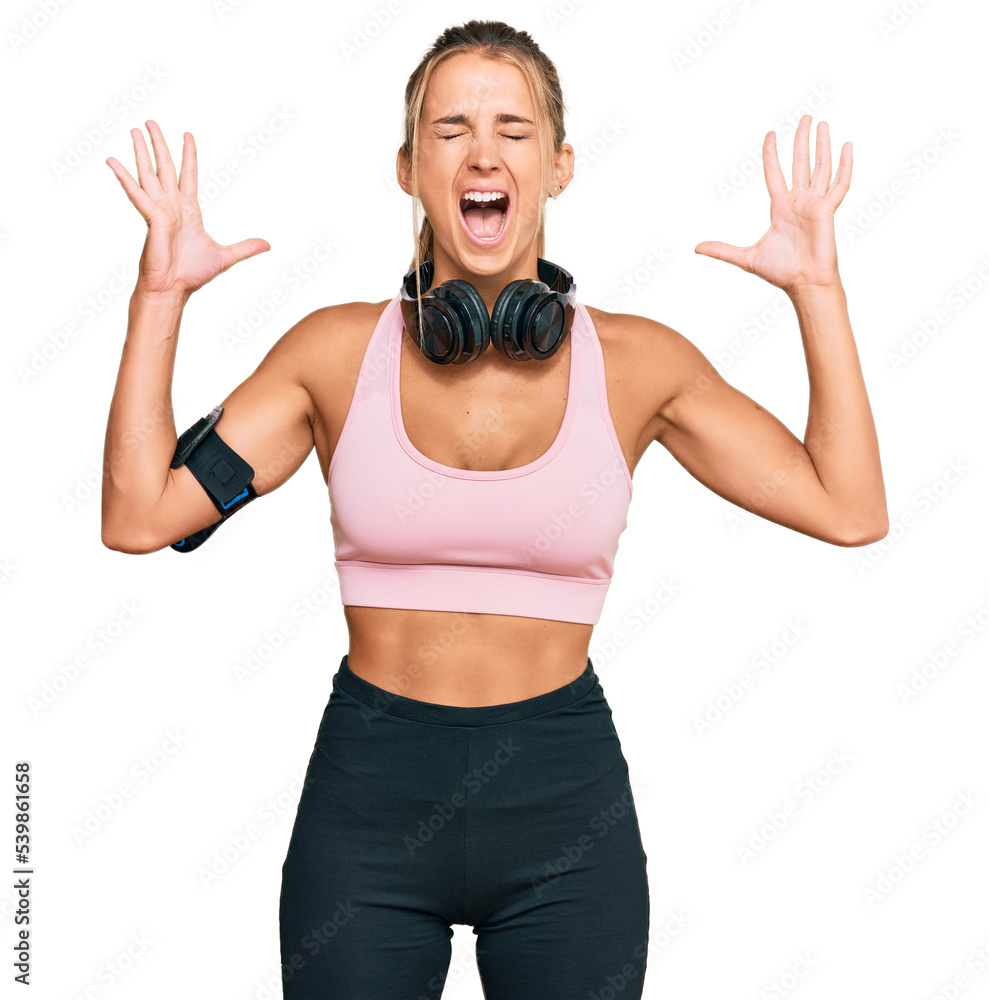 Young blonde woman wearing gym clothes and using headphones celebrating mad and crazy for success with arms raised and closed eyes screaming excited. winner concept Young blonde woman wearing gym clothes and using headphones celebrating mad and crazy for success with arms raised and closed eyes screaming excited. winner concept